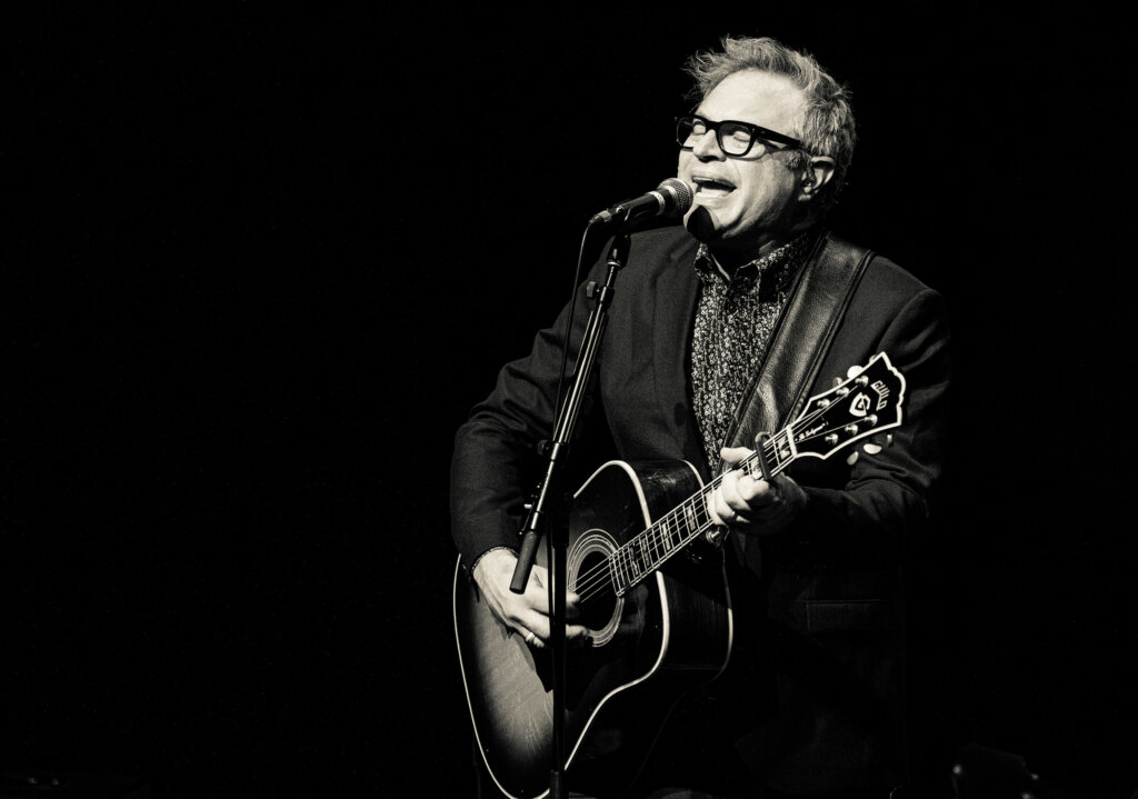 Steven Page sings into a microphone while playing guitar during a musical performance.