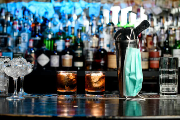 PPE mask hangs on cocktail shaker next to two mixed drinks on a bartop.